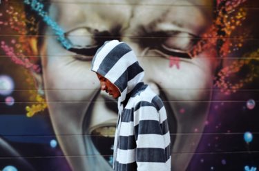 man wearing black and white striped hoodie standing near wall with graffiti of woman's face singing