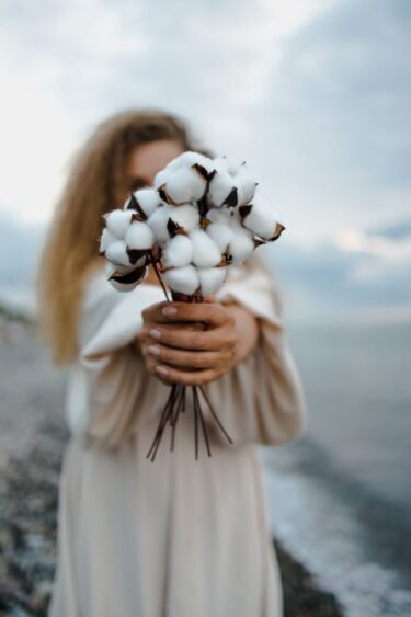 A woman holds a bouquet of cotton flowers with a blurred seaside backdrop, capturing a serene moment.